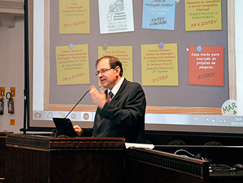 Foto em plano médio traz o desembargador Célio Horst Waldraff, posicionado atrás do púlpito do Plenário, falando ao público. Ao fundo, parte da tela de projeção com imagem do calendário de eventos da Escola Judicial.