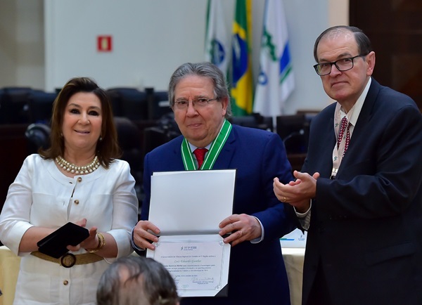 Imagem: Fotografia de dois homens e uma mulher em pé, uma ao lado da outra, posam para a fotografia. Da esquerda para a direita, a desembargadora Ana Carolina Zaina, veste casaco branco e usa colares; desembargador Luiz Eduardo Gunther, veste terno azul co gravata vermelha, ele tem em mão um diploma e um cola com a homenagem, com fita verde e branca; o desembargador presidente do TRT-PR, Célio Waladraff, de terno azul com gravata vermelha e branca, ele aplaude o momento. (arquivo JPG)