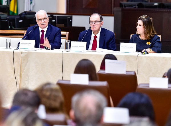 Imagem; fotografia: Ministro Edson Fachin fala sentado em frente a uma mesa com toalha creme. Ao lado, o presidente do TRT-PR, desembargador Célio Waldraff, e a diretora da Escola Judicial, desembargadora Ana Carolina Zaina. Em frente deles, uma plateia acompanha o discurso. (arquivo JPG)