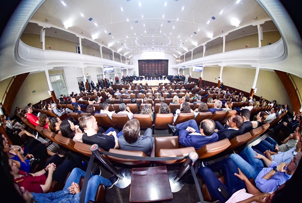 Imagem: Fotografia com lente olho de peixe (que distorce as bordas) mostra o plenário Pedro Ribeiro Tavares lotado com a plateia de costas. Ao fundo, a mesa de autoridades na abertura do evento. (arquivo JPG)